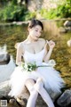 A woman in a white dress sitting on a rock by a river.